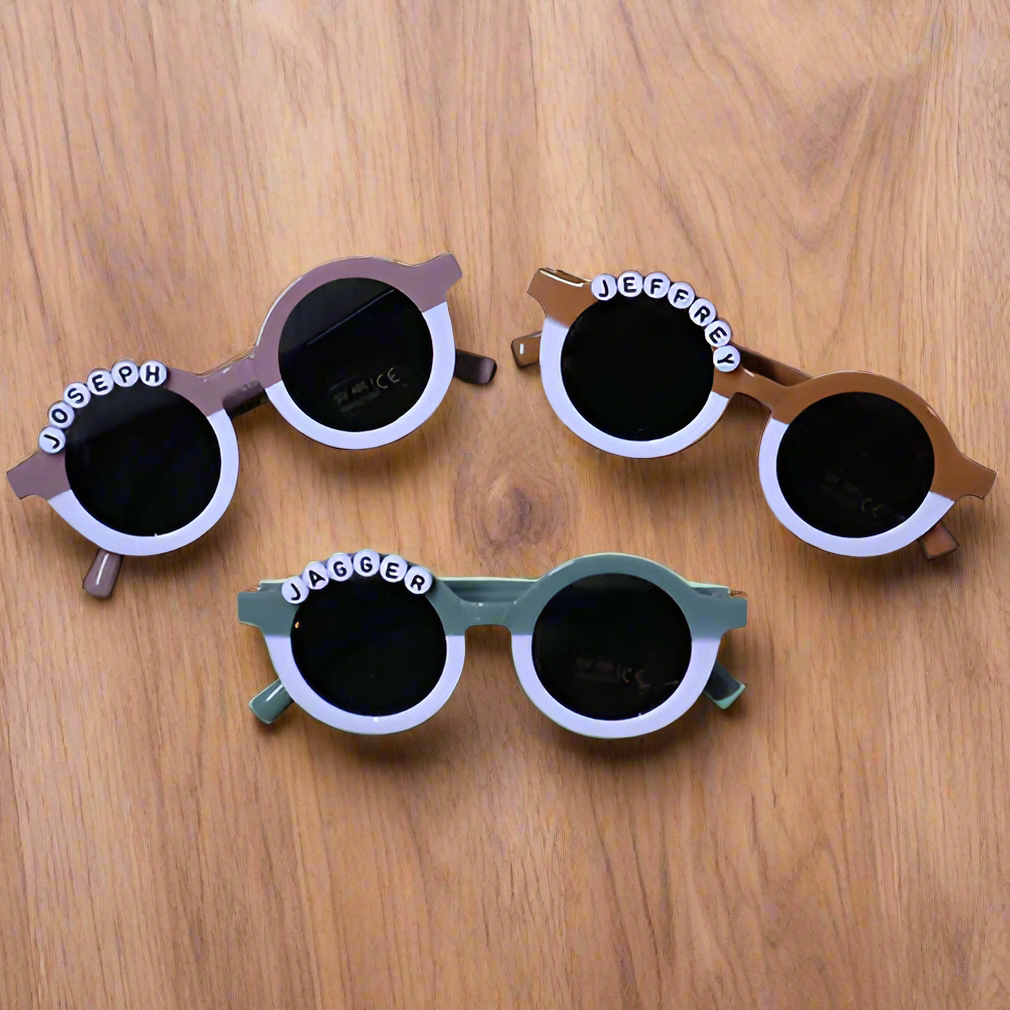 Three pairs of children's sunglasses with personalized names on a wooden surface