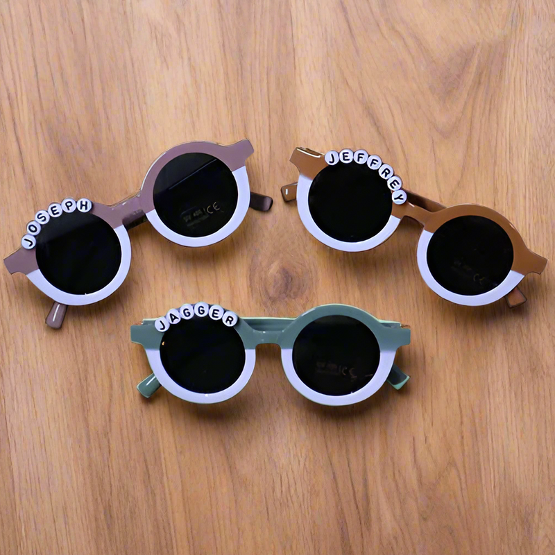 Three pairs of children's sunglasses with personalized names on a wooden surface